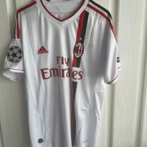 AC Milan Pato 7 Champions League tröja - Snygg AC Milan bortatröja från Adidas med Pato och nummer 7 på ryggen. Vit bas med röda och svarta detaljer, Champions League-märke på ärmen och Fly Emirates-tryck på bröstet. Klassisk V-ringning och korta ärmar. Tillverkad i lätt funktionsmaterial.