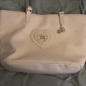 Ljusrosa axelväska Juicy Couture - Superfin ljusrosa axelväska från Juicy Couture med präglade JC-loggor och hjärtformad detalj framtill. Väskan har guldfärgade metalldetaljer, dragkedja upptill och ett innerfoder med Juicy Couture-tryck. Perfekt storlek för allt du behöver ha med dig. Väskan är köpt i usa för cirka 3000 svenska kronor och är i bra skick 