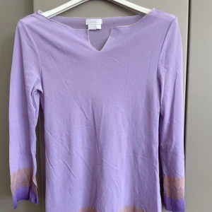 Ljuslila långärmad blus med mönster - Ljuslila blus från Zara med lång ärm och v-ringning. Blusen har dekorativa guldfärgade mönster längs ärmslut och nederkant, vilket ger en unik touch. Tillverkad i mjukt bomullstyg, perfekt för dig som gillar färg och detaljer. S/M 
