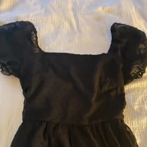 Svart kortklänning med puffärm - Supersöt svart kortklänning med transparenta puffärmar och diskret prickigt mönster. Klänningen har en rak urringning och lätt volym i kjolen samt knytning baktill. Perfekt för dig som gillar en trendig och lekfull stil.