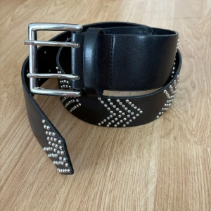 Svart skärp med nitar och dubbla spännen - Snyggt svart bälte i skinn med bred design och coola silvriga nitar i pilmönster längs hela bandet. Bältet har ett rejält dubbelt spänne i silverfärg som ger en edgy vibe. Perfekt accessoar för att lyfta din outfit.