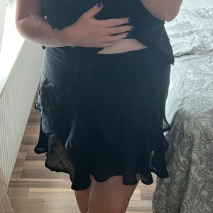 Svart volangkjol med omlottkänsla - Supersöt svart kortkjol med volangkant och lätt omlottkänsla. Kjolen har en flowy passform och är perfekt för dig som gillar en feminin och trendig stil. Materialet känns lätt och luftigt, vilket gör den skön att bära under varma dagar.