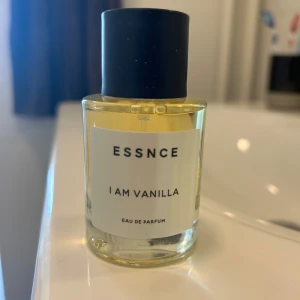 ESSNCE I Am Vanilla parfym - En stilren parfymflaska från ESSNCE med doften I Am Vanilla. Flaskan är genomskinlig med ljusgul vätska och har en svart, matt kork. Minimalistisk vit etikett med svart text. Perfekt för dig som älskar söta och varma vaniljtoner.