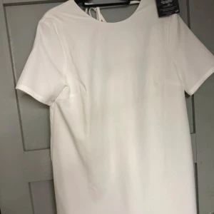 Vit kortklänning med knyt i ryggen - En vit kortklänning från Nelly med korta ärmar och v-ringning i ryggen med knytband. Klänningen har en rak passform. Aldrig använd.