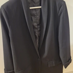 Svart klassisk kavaj - Stilren svart kavaj med enkel knäppning och klassiska slag. Kavajen har två fickor framtill och är helfodrad. Perfekt för en clean och tidlös look. Materialet känns mjukt och har en lätt glans.