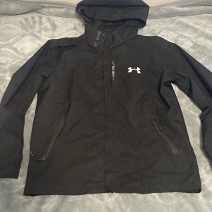 Svart vindjacka från Under Armour M - Svart vindjacka från Under Armour med huva och dragkedja. Jackan har vita logodetaljer på bröstet och texten 'Explore' framtill. Två fickor med dragkedja och ventilerande meshfoder. Perfekt för aktiva dagar när du vill hålla stilen.