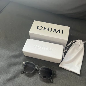 Svarta solglasögon från CHIMI - Snygga solglasögon från CHIMI med transparent svart/grå båge och runda, mörka linser. Kommer med originalfodral och tygpåse. Perfekt accessoar för att lyfta din stil och skydda ögonen mot solen.