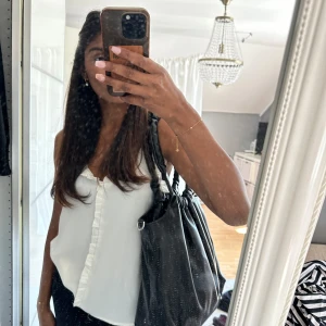 Svart axelväska i skinnimitation - Svart axelväska med scrunchie-inspirerade handtag och dragsko upptill. Väskan är rymlig och har en innerficka med dragkedja. Tillverkad i mjuk skinnimitation med snygg glansig finish. Materialet på handtaget har lossnat lite, men ingeting man ser när man dragit ned materialet. 