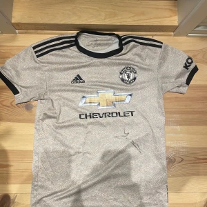 Manchester United bortatröja Adidas - Snygg Manchester United bortatröja från Adidas med beige bas och subtilt mönster. Lite skadat Chevrolet-tryck på bröstet. Kommer tyvärr inte ihåg vem som signerat tröjan! Priset går att diskutera pågrund av den lilla skadan