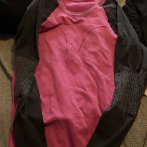 Rosa och svart långärmad träningströja - Sportig långärmad träningströja i rosa och svart med åtsittande passform. Tröjan har rund halsringning och är tillverkad i ett stretchigt syntetmaterial som andas, perfekt för aktiva dagar. Snygga kontrastpaneler längs sidorna och ärmarna. Står ej storlek men gissar på S 