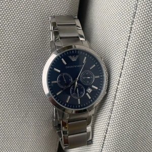 Emporio Armani silverfärgad klocka - Stilren klocka från Emporio Armani med blå urtavla och silverfärgat armband i metall. Klockan har tre mindre urtavlor, datumvisning och klassisk länkdesign. Perfekt accessoar för dig som gillar en clean och modern look.