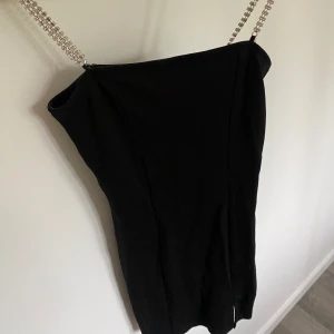 Svart kort klänning med glittriga band - Supersnygg svart kort klänning med smala axelband i glittrande strass. Enkel och stilren design som sitter tight och markerar figuren. Perfekt för dig som vill sticka ut med en elegant men ändå cool look.