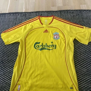 Liverpool gul bortatröja Adidas - Liverpool FC:s klassiska gula bortatröja från Adidas med röda detaljer och Carlsberg-logga på bröstet. Tröjan har klubbmärket broderat och tre röda ränder längs axlarna. Tillverkad i lätt och ventilerande material, perfekt för match eller samling. Pris går att diskutera. 