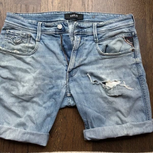Replay ljusblå jeansshorts herr - Snygga ljusblå jeansshorts från Replay med slitna detaljer och upprullade benslut. Klassisk femficksmodell med dragkedja och knapp. Perfekta för sommaren och ger en avslappnad vibe.