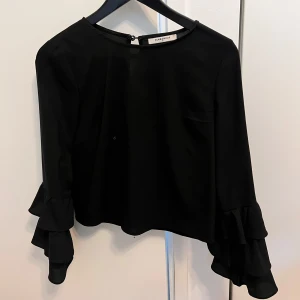 Svart blus med volangärmar Glamorous - Supersnygg svart blus från Glamorous i storlek XS. Blusen har långa ärmar med dubbla volanger som ger en extra cool touch. Rak passform och rund halsringning. Perfekt att styla med jeans eller kjol för en trendig look.