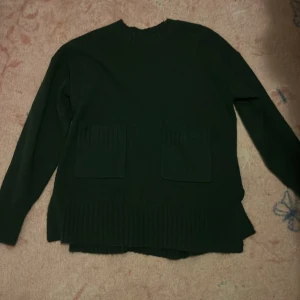 Grön stickad tröja från KappAhl - Mörkgrön stickad tröja från KappAhl i storlek S. Tröjan har två stora fickor framtill, ribbade detaljer vid nederkant och långa ärmar.
