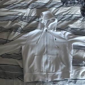 Vit hoodie från Polo Ralph Lauren SIZE M - Snygg vit hoodie från Polo Ralph Lauren med dragkedja och klassisk logga på bröstet. Tröjan har huva med snörning, två fickor fram och är tillverkad i mjuk bomullsmix. Perfekt för en clean och stilren look.