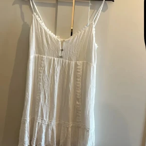 Vit somrig klänning Pull&Bear S - Luftig vit kortklänning från Pull&Bear i storlek S. Klänningen har tunna axelband, v-ringning med små knappar framtill och volangkant nertill. Tillverkad i bomull med broderade detaljer längs kanterna. Perfekt för varma dagar och en chill stil.