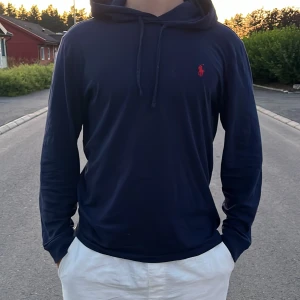 Marinblå hoodie från Ralph Lauren - Snygg marinblå hoodie från Ralph Lauren med röd logga på bröstet. Tröjan har huva med snörning och långa ärmar. Perfekt för dig som gillar en stilren och klassisk look.