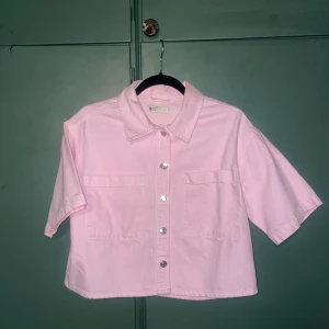 Ljusrosa jeansskjorta från Gina Tricot - Kortärmad jeansskjorta i ljusrosa från Gina Tricot. Modellen är boxig och croppad med två stora bröstfickor och silvriga knappar framtill. Använd 1 gång.