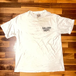 Vit t-shirt från Gallery Dept. - Vit t-shirt från Gallery Dept. med svart tryck fram och stort rött tryck bak där det står 'GALLERY DEPT. HOLLYWOOD, CA.'. Klassisk passform och rund halsringning. Perfekt för dig som gillar streetwear och enkel stil.