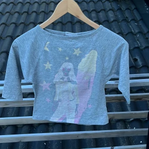 Grå croppad topp med tryck Lindex - Grå croppad topp från Lindex Girls med coolt tryck av två figurer och stjärnor i pastellfärger på framsidan. Toppen har trekvartsärm och bred halsringning. Perfekt för dig som gillar unika prints och en avslappnad stil.