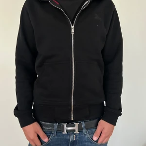 Svart hoodiejacka från Burberry M - Svart hoodiejacka från Burberry i storlek M. Jackan har hel dragkedja, huva och klassiskt rutigt Burberry-foder på insidan. Tillverkad i mjuk bomull, med ribbade muddar och fickor framtill. Perfekt för en clean och stilren look. Har tagit av snörena men dom följer med vid köp 