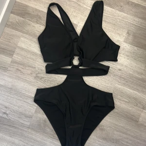 Svart cut out bikini med ringdetalj - Snygg svart bikini med högt skurna trosor och trendig cut out-design. Den har breda axelband och en cool ringdetalj framtill som ger en unik look. Gjord i stretchigt syntetmaterial för bästa passform. Aldrig använd