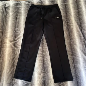 Svarta mjukisbyxor från Les Deux - Svarta mjukisbyxor från Les Deux med raka ben och diskret logga på ena sidan. Byxorna har dragsko i midjan och är tillverkade i ett mjukt, slätt material som känns riktigt skönt. Perfekta för en chill och stilren look.