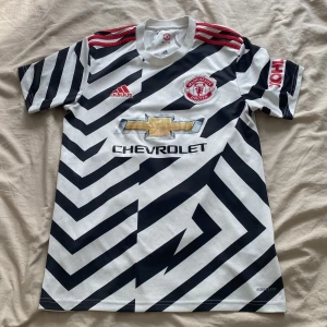 Manchester United fotbollströja  - Säljer en Manchester United bortatröja från Adidas med svartvitt zebramönster, röda axelränder och klubbmärke på bröstet. Tröjan har Chevrolet-logga framtill och är tillverkad i lätt funktionsmaterial. Perfekt för dig som vill sticka ut på planen eller läktaren.