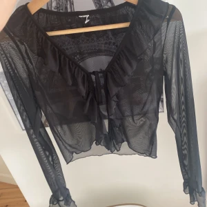 Svart mesh blus med volang - Säljer en svart meshblus med långa ärmar då den är för stor, volangdetaljer längs v-ringningen och ärmsluten samt knyt framtill. Blusen är croppad och har en riktigt snygg och trendig look som passar perfekt till ett par jeans eller något annat. Stl 158/164/xs.