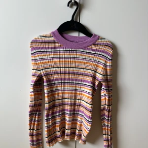 Randig ribbad tröja från Lindex XS - Superfin långärmad tröja från Lindex i ribbat material. Färgglada ränder i lila, orange, svart och vitt med en bred lila krage. Sitter tajt och är perfekt för dig som gillar retrovibbar och vill sticka ut lite extra.