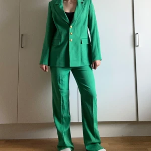 Grön kostym från Yosoy med guldknappar - Stilren grön kostym från Yosoy med kavaj och raka byxor. Kavajen har snygga guldknappar och klassiska slag, samt fickor med lock. Byxorna har matchande knappdetalj i guld. Kostymen har en modern, rak passform och är tillverkad i ett mjukt, lätt material.