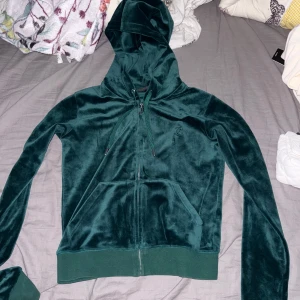 Juicy Couture grön velour hoodie - Mörkgrön hoodie i mjuk velour från Juicy Couture. Jackan har huva, dragkedja framtill, snörning vid halsen och känguruficka. Perfekt för en chill och trendig look, med diskret JC-logo broderad på bröstet. Matcha med byxorna för en komplett tracksuit.