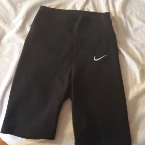 Svarta ribbade cykelshorts Nike - Svarta ribbade cykelshorts från Nike med hög midja och klassisk vit swoosh-logga på låret. Stretchigt material som sitter tight och bekvämt, perfekta för träning eller chill. Snygg och enkel design som funkar till det mesta.