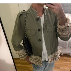 Grön jacka med spetsdetaljer Zara Woman - Snygg militärgrön jacka från Zara Woman med unika virkade spetsdetaljer och tofsar i beige längs ärmslut och nederkant. Jackan har två bröstfickor, stora knappar och rak passform. 