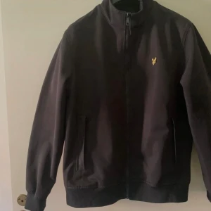 Svart bomberjacka från Lyle & Scott - Svart bomberjacka från Lyle & Scott med klassisk gul logga på bröstet. Jackan har dragkedja framtill, ribbade muddar och två fickor. Snygg och stilren modell som passar till det mesta. Materialet är troligtvis polyester. Finns ett litet hål bak på jackan som synes på en bild . Annars väldigt lite andvänd 