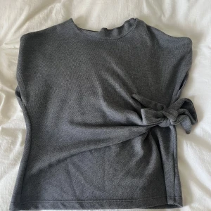 Grå stickad topp med knyt från Zara - Mörkgrå stickad topp från Zara i storlek M. Modellen är ärmlös med rund hals och har en snygg knytdetalj i sidan som ger en cool twist. kan inte styla den på rätt sätt så har tyvärr bara använt 1 gång och aldrig mer 🤍