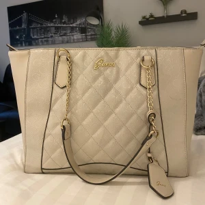 Beige quiltad handväska från Guess - Snygg beige handväska från Guess med quiltad framsida, guldfärgade kedjedetaljer och logotyp. Väskan har flera innerfack och dragkedja upptill. Materialet är syntet med mönstrad insida och klassisk Guess-tag.