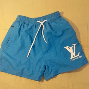 Blå Louis Vuitton badshorts med snörning - Blåa badshorts från Louis Vuitton med vit LV-logga på ena benet. Shortsen har elastisk midja och vit snörning framtill för perfekt passform. Tillverkade i lätt syntetmaterial som torkar snabbt, perfekta för stranden eller poolen.
