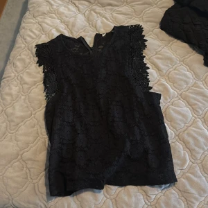 Svart spetsblus med blommiga detaljer - Supersnygg svart blus i spets med blommigt mönster och coola virkade detaljer vid axlarna. Ärmlös modell med lätt v-ringning och rak passform. Perfekt för dig som gillar en stilren men ändå unik look. Pris kan diskuteras 