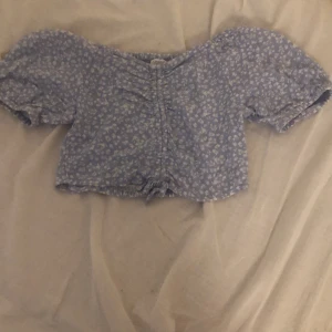 Blommig croppad topp från Lindex - Söt ljusblå croppad topp från Lindex med vita småblommor. Toppen har korta puffärmar, offshoulder-modell och smockad rygg för extra stretch. Framtill finns snörning och rynkade detaljer. Perfekt för en somrig look!