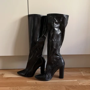 Svarta höga stövlar med klack - Snygga svarta stövlar med högt skaft och chunky klack. Skorna har ett croco-mönstrat konstläder som ger en cool och trendig vibe. Spetsig tå och plattformsinspirerad sula. Perfekta för att lyfta vilken outfit som helst.