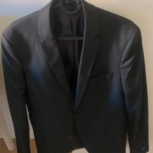 Svart kavaj Jack & Jones Premium 46 - Stilren svart kavaj från Jack & Jones Premium i slim fit-modell. Klassisk design med två knappar framtill, smala slag och fyra dekorativa knappar vid ärmslutet. Perfekt för dig som vill ha en snygg och modern look.
