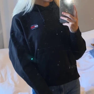 Svart hoodie - Svart hoodie från Tommy Jeans med klassisk logga på bröstet. Tröjan har huva med snörning och en stor magficka framtill. 