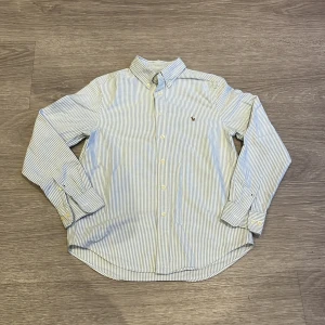 Randig skjorta från Ralph Lauren - Snygg skjorta från Ralph Lauren i vitt och blått med tunna ränder. Klassisk button-down krage, broderad logga på bröstet och långa ärmar med manschett. Skjortan är i bomull och har en normal passform. Perfekt för dig som gillar stilrena och tidlösa plagg.