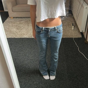 Blå low waist bootcut jeans från Gina tricot - Säljer ett par blå jeans med låg midja och bootcut-snitt. Jeansen har snygga detaljerade bakfickor med knappar och dragkedjor. Klassisk femficksmodell i mjukt denimtyg, perfekt för dig som gillar en avslappnad men trendig look.