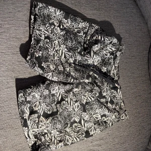 Svarta blommiga badshorts med snörning - Snygga svarta badshorts med vitt blommigt mönster och praktisk snörning i midjan. Ficka på höger ben med dragkedja. Perfekta för sommardagar när du vill sticka ut lite extra. Lätta och bekväma med avslappnad passform.