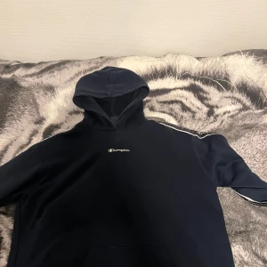 Svart Champion hoodie med vita detaljer - Svart hoodie från Champion i storlek small. Klassisk modell med huva, vita ränder längs axlarna och broderad Champion-logga på bröstet. Mjuk bomullskänsla och känguruficka framtill. Perfekt för en chill och sportig look. Den är använd nån gång annars har den bara stått i garderoben, vill bli av med den bara.
