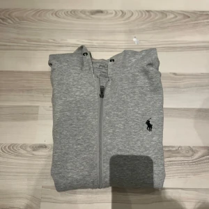 Grå hoodie från Polo Ralph Lauren - Snygg grå hoodie från Polo Ralph Lauren med dragkedja och klassisk huva. Tröjan har det ikoniska broderade logotypen på bröstet och är tillverkad i mjuk bomullsmix. Perfekt för chill dagar eller när du vill ha en clean och stilren look.